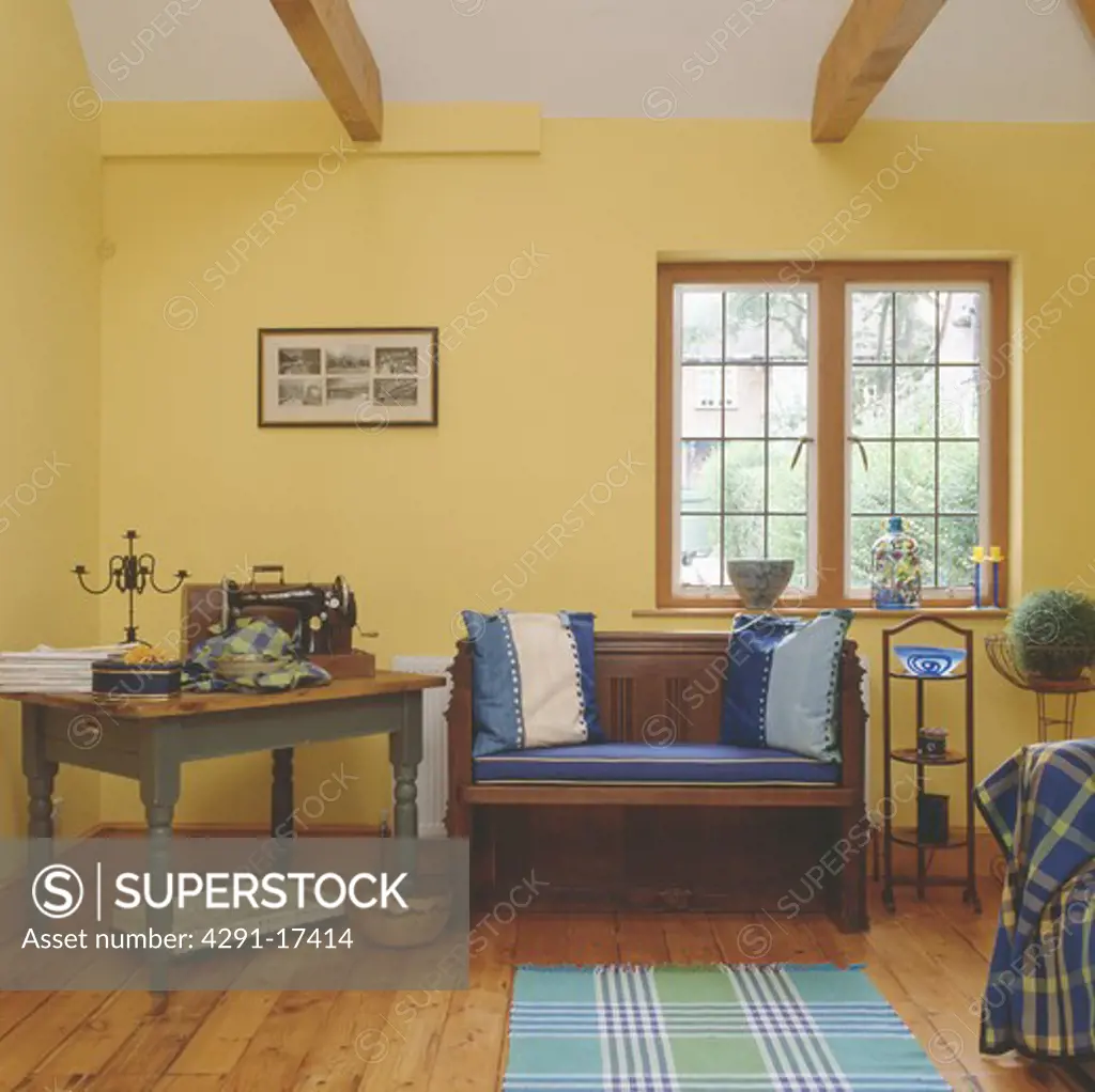 Sewing machine on table beside wooden settle with blue cushions below window in yellow living and workroom