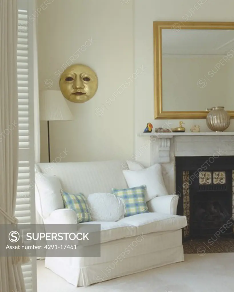 White and checked cushions on small white sofa beside fireplace in white living room with gilt sun sculpture on wall
