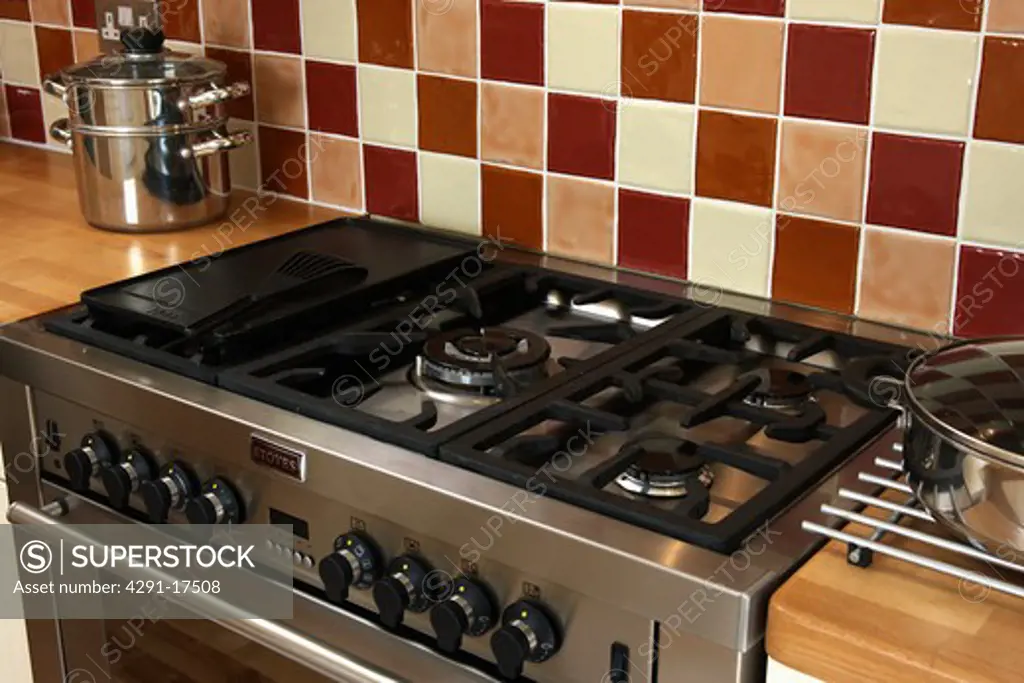 Close-up of gas hob on stainless steel range oven below brown and cream tiled wall