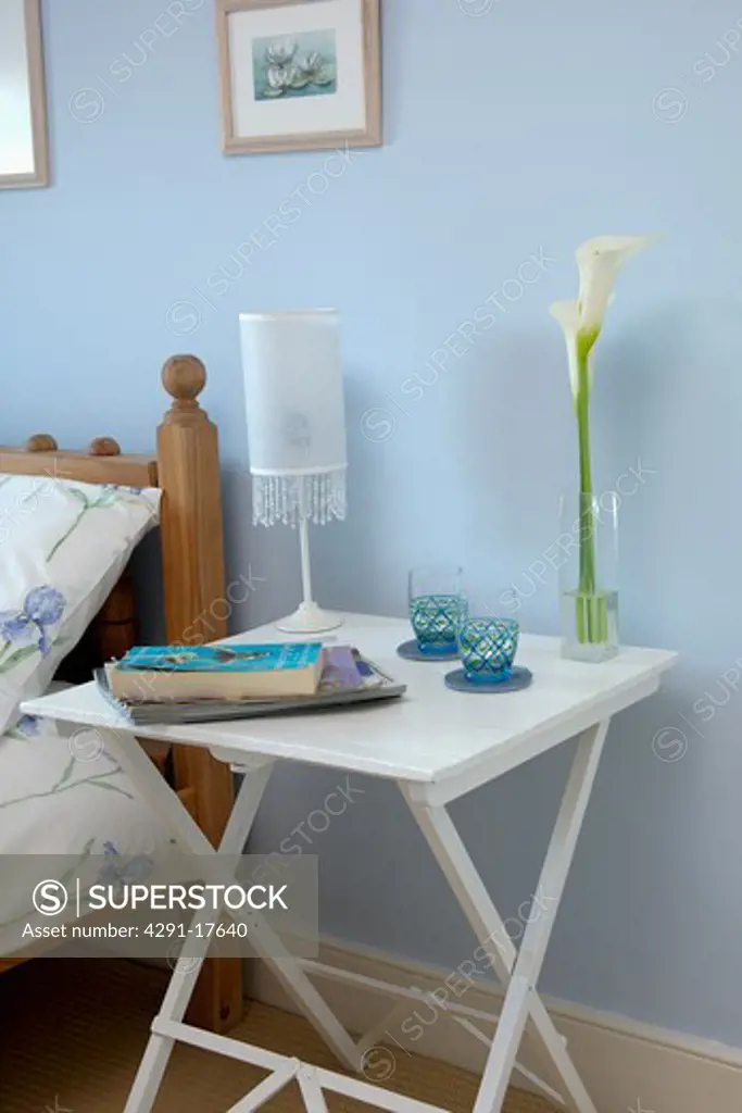 Lamp and books on small white painted bedside table in pastel blue bedroom