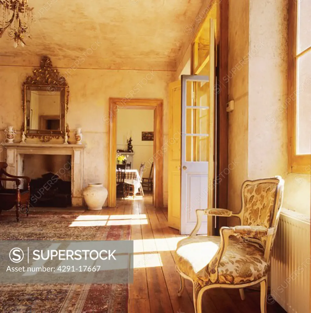 Antique chair and rough-plastered walls in country hall with large mirror above fireplace and sunlight on floor