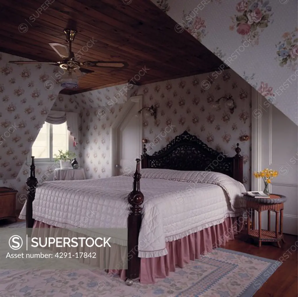 White quilt and pink vallance on carved mahogany bed with turned posts in attic bedroom with floral wallpaper