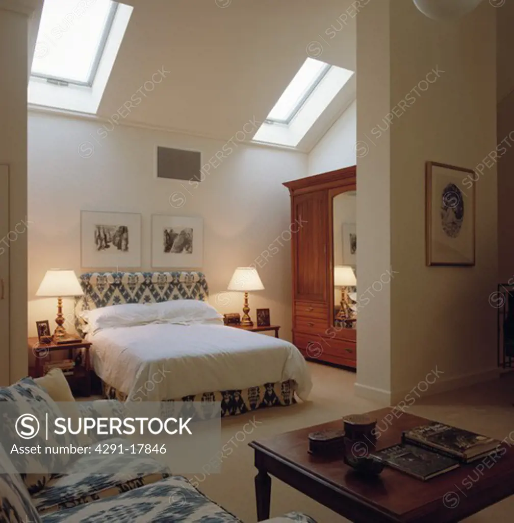 Lighted lamps on either side of blue and white upholstered bed in modern white bedroom extension with Velux windows