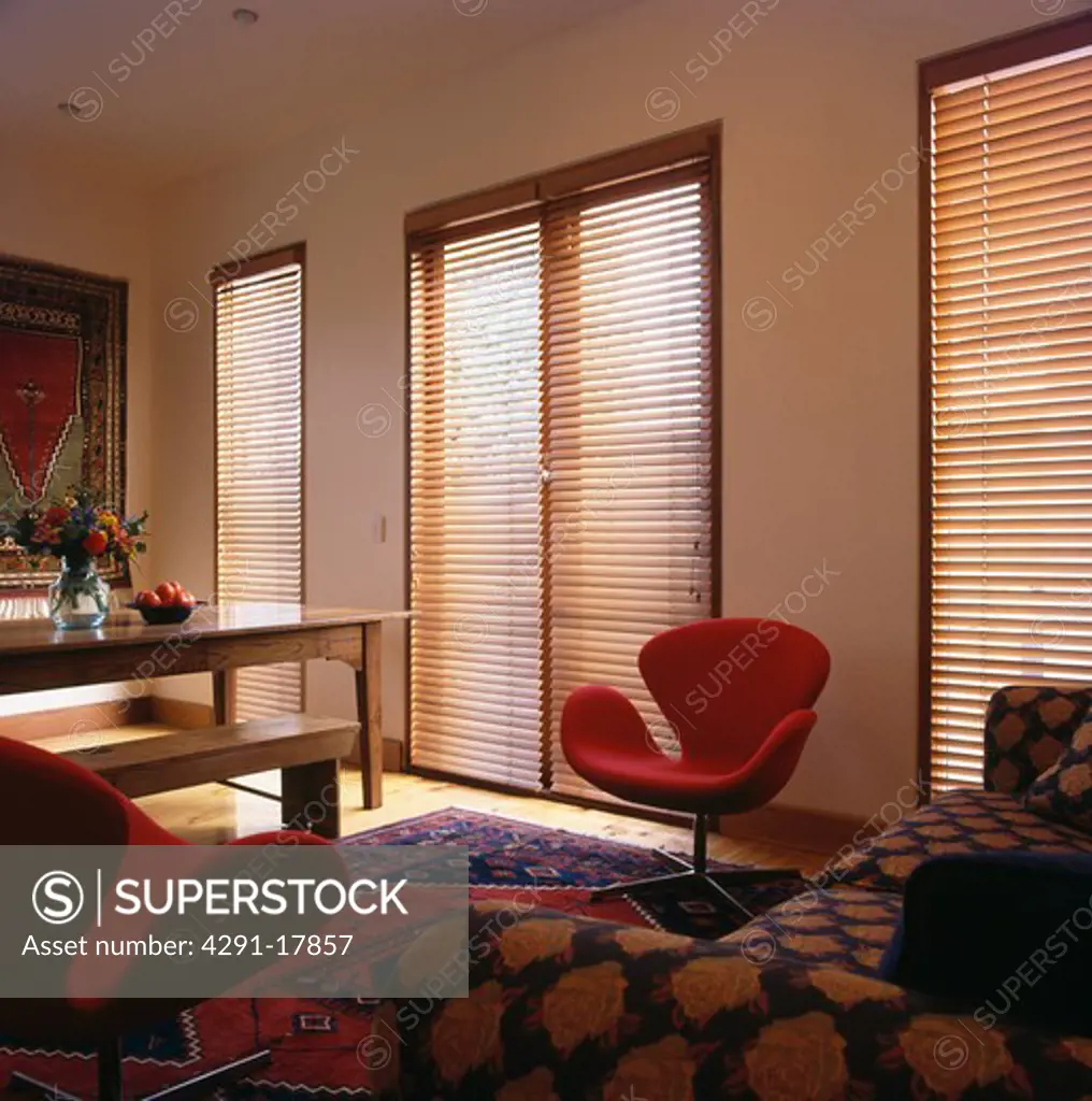 Slatted wooden blinds on tall windows in modern living room with red Swan chair