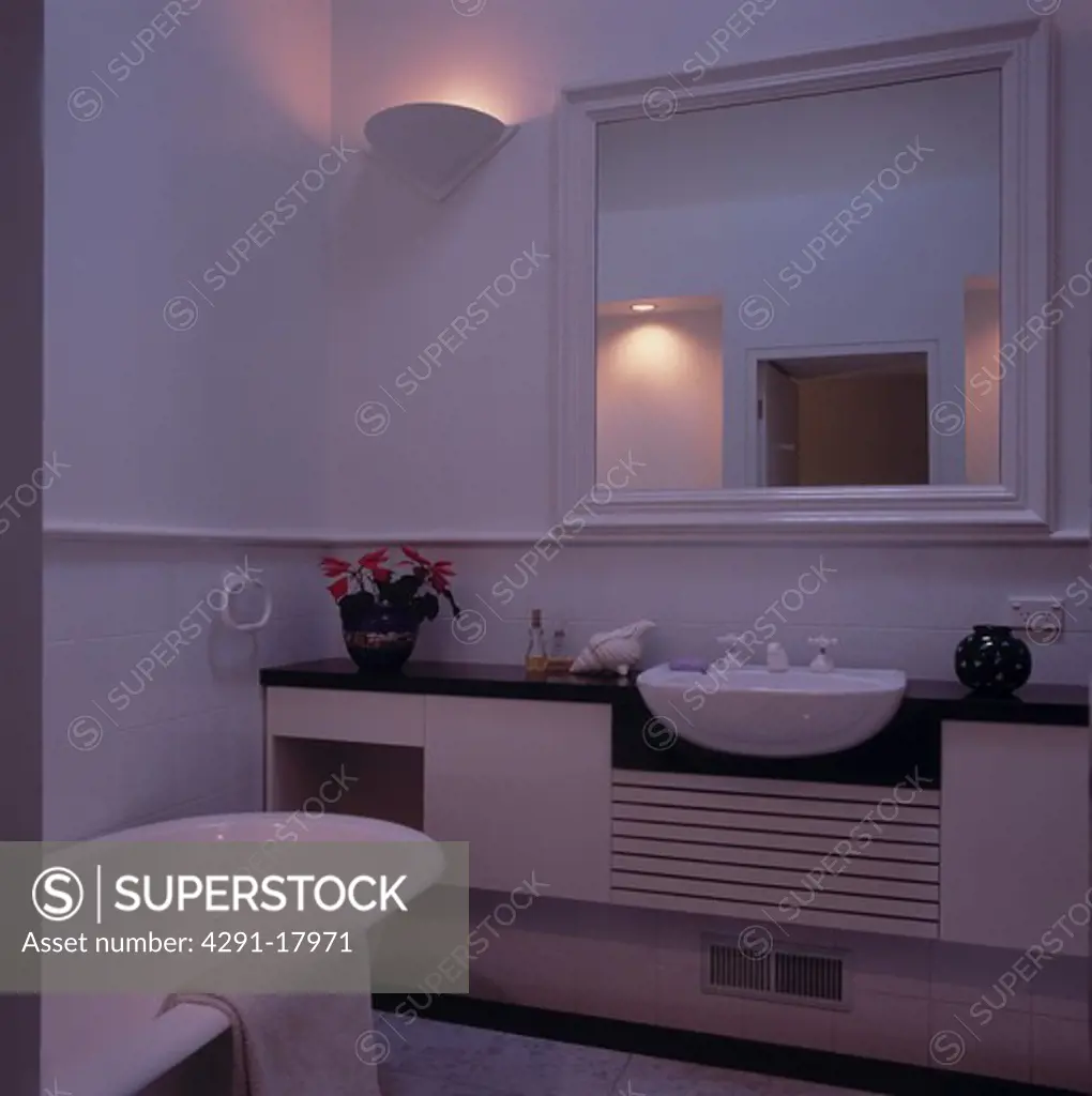 Rectangular white mirror above basin in vanity unit in modern white bathroom with wall uplighter