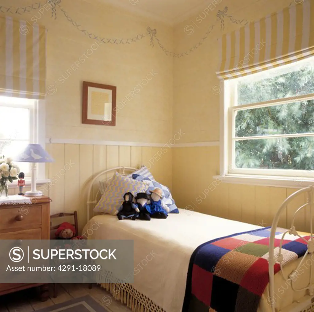 Yellow striped blinds at window above white wrought iron single bed with knitted patchwork blanket and cream throw