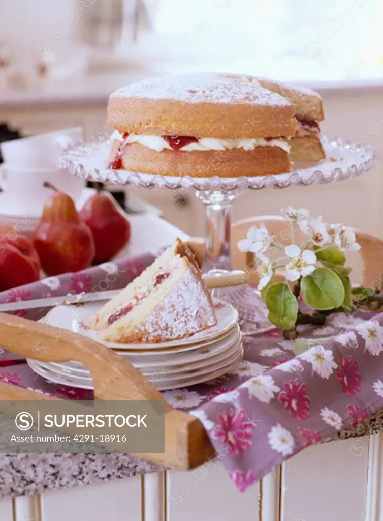 Slice of cake on pile of plates in front of spongecake on glass cakestand