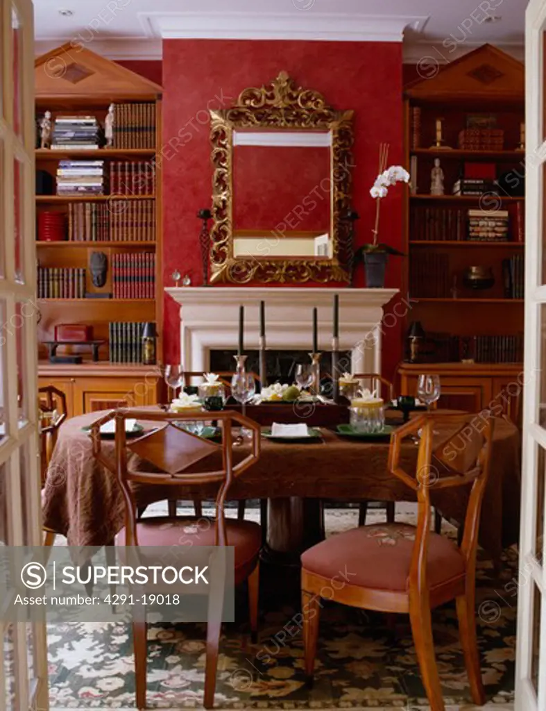 Doors open to Biedermeier chairs in red dining room with tall bookcases on either side of fireplace below ornate gilt mirror