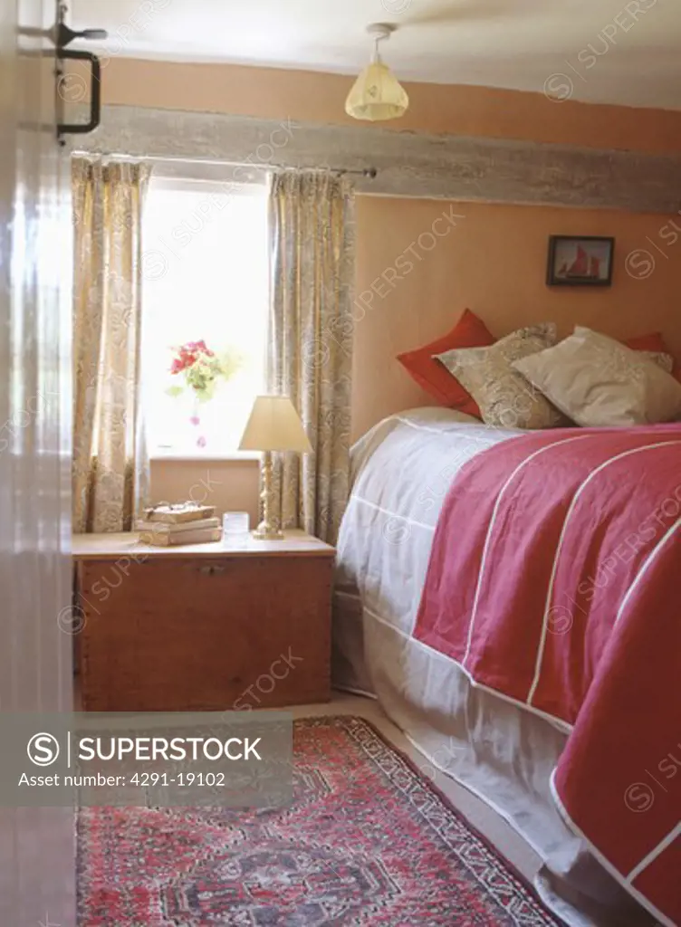 Wooden chest below window in cottage bedroom with cushions and red throw on the bed
