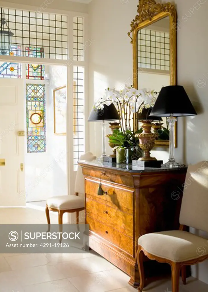 Gilt mirror above antique chest-of-drawers with black lamps in traditional white hall with limestone floor tiles