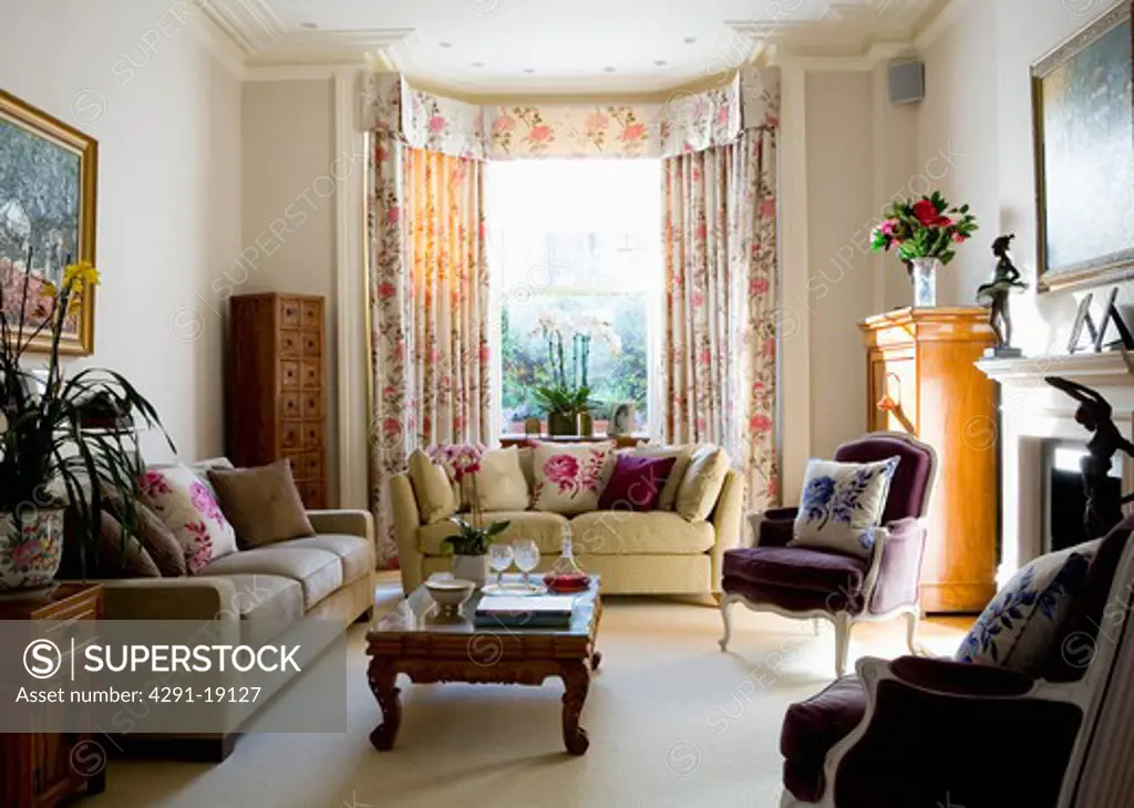 Cream sofas and purple velour French-style armchairs in living room with cream carpet and floral curtains
