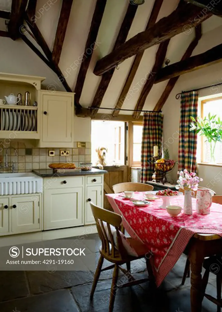 Red floral cloth on table in cream country kitchen with stone flooring and beamed apex ceiling