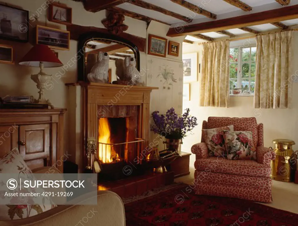 China Staffordshire dogs on wooden mantelpiece above fireplace with lighted fire in cottage living room with red patterned armchair