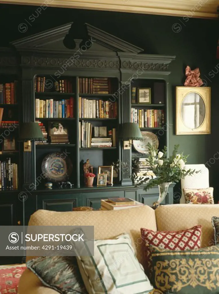 Fitted bookshelves with pediment in dark green living room with cushions piled on cream sofa