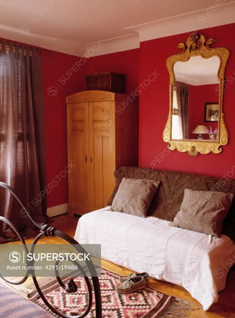 Ornate gilt mirror above couch with brown cushions and cream throw in red bedroom with pine wardrobe