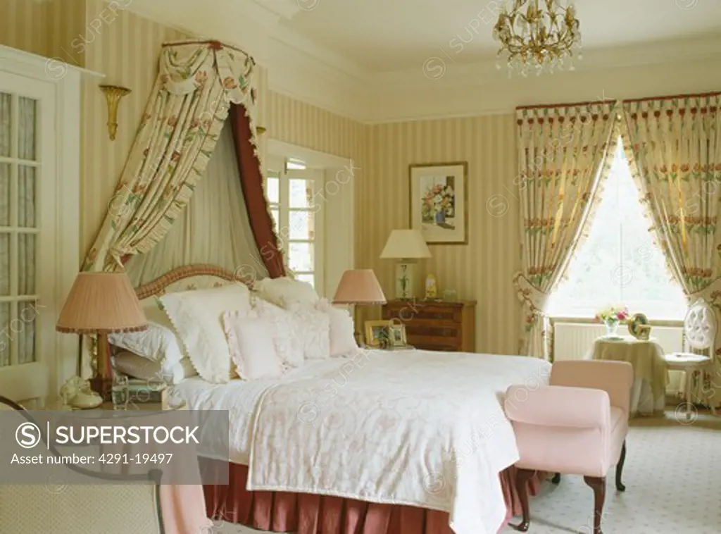 Coronet with floral drapes above bed piled with cushions in country bedroom with striped wallpaper and floral curtains