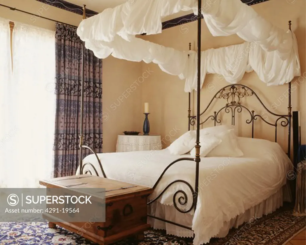 Antique wooden chest below black wrought iron four-poster bed with white bed linen and draped white cotton on rails
