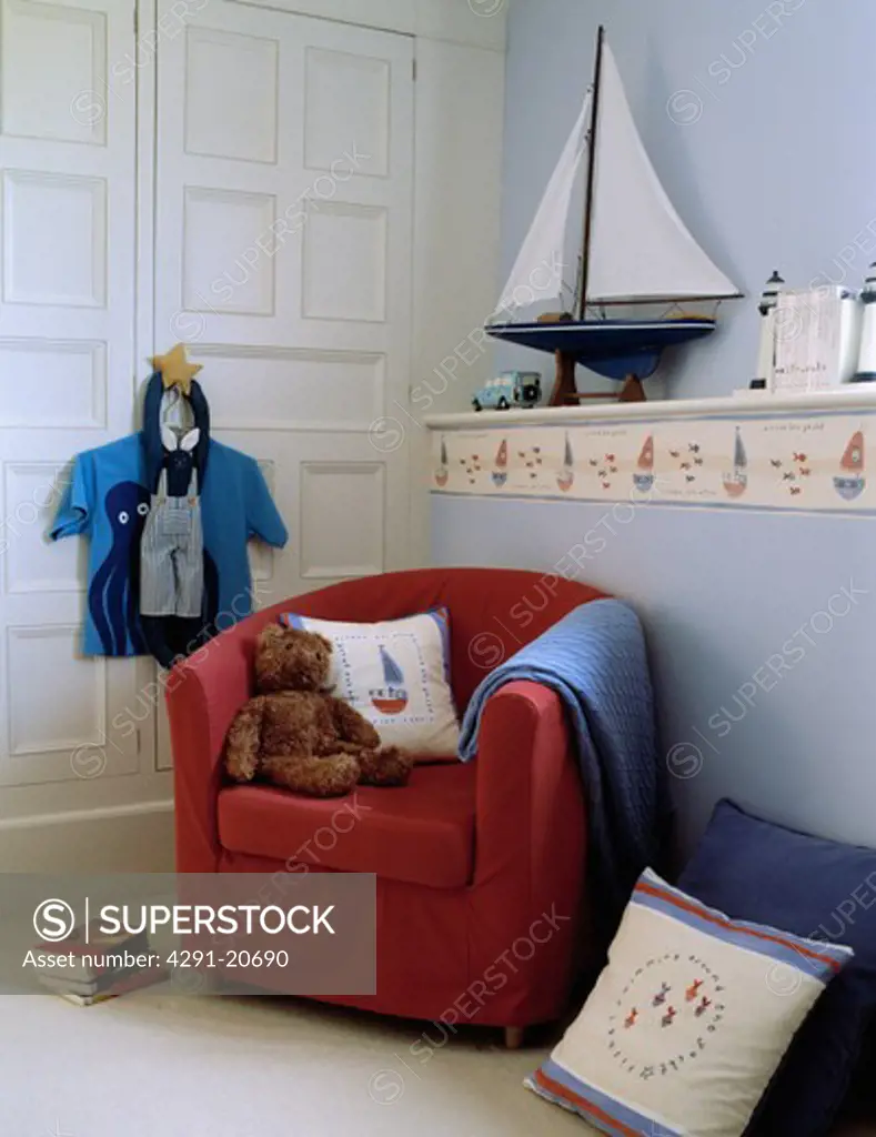 Teddybear with cushion and blue throw on red tub armchair in nautical-themed child's bedroom with sailing boat on shelf