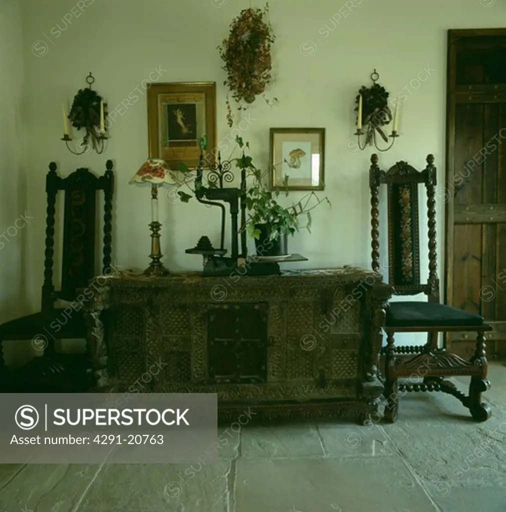 Antique Jacobean-style chairs on either side of carved wooden cupboard in country hall with flagstoned floor