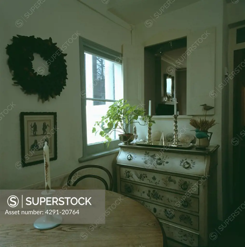 Candle on circular pine table in small dining room with floral painted bureau beside window