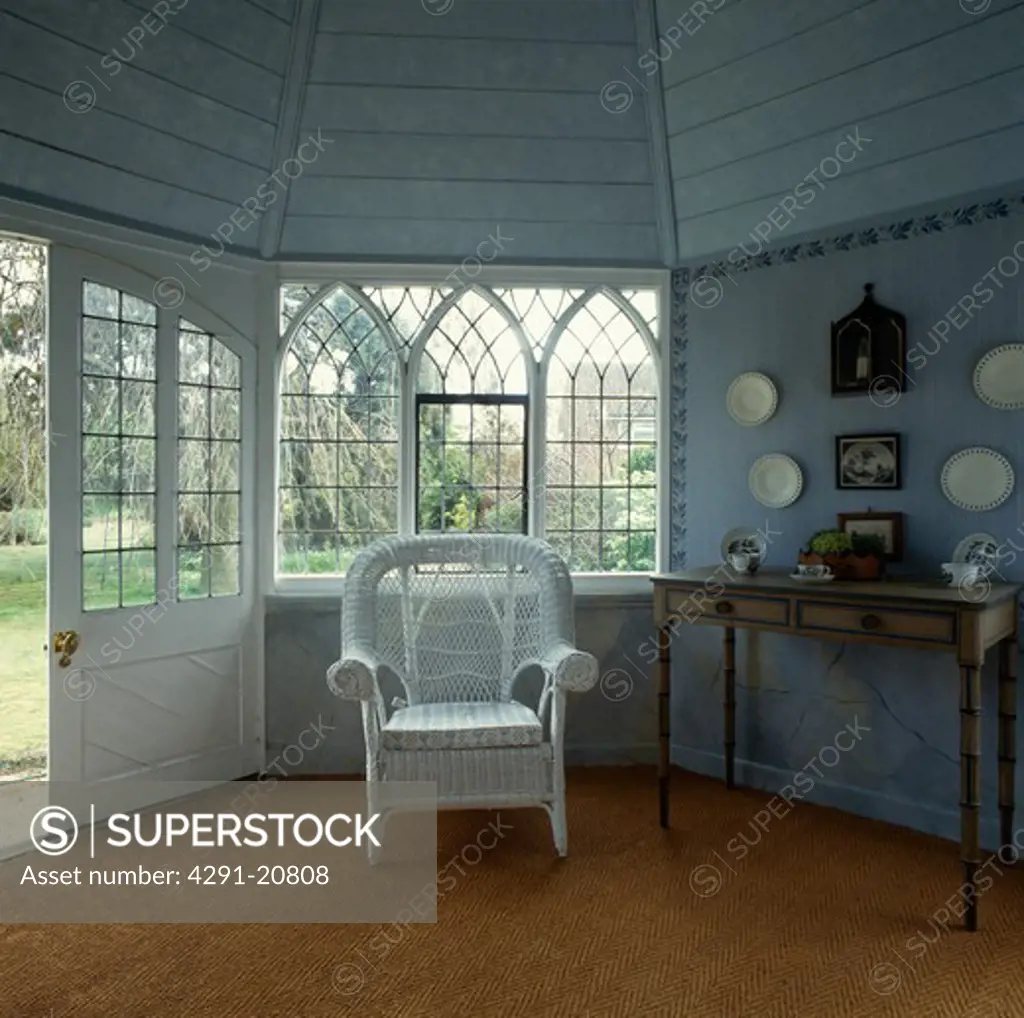 White wicker armchair in front of Gothic-style window in pastel blue hall with sisal carpet and wooden ceiling