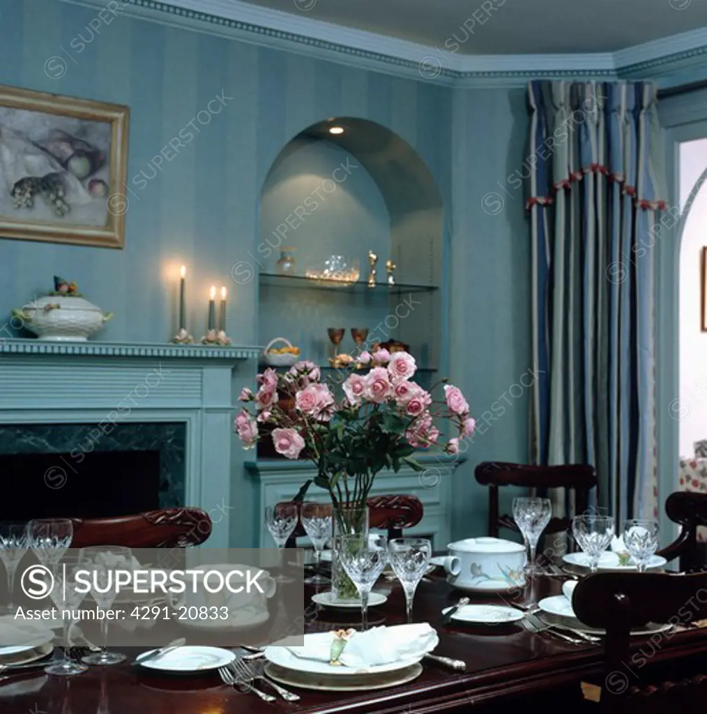 Pinks in glass vase on table with place settings in pale blue dining room with lighted shelving in alcove