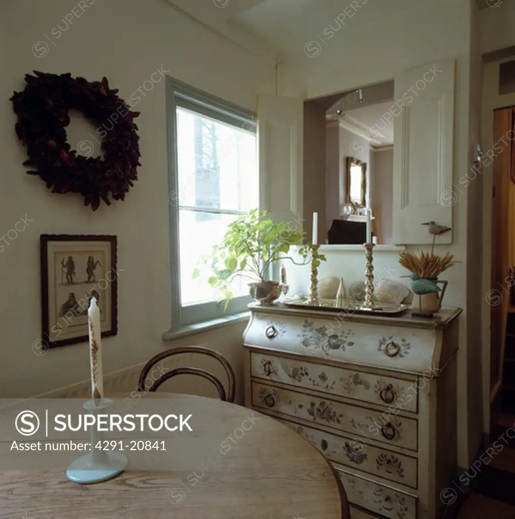 Candle on circular pine table in small dining room with floral painted bureau beside window