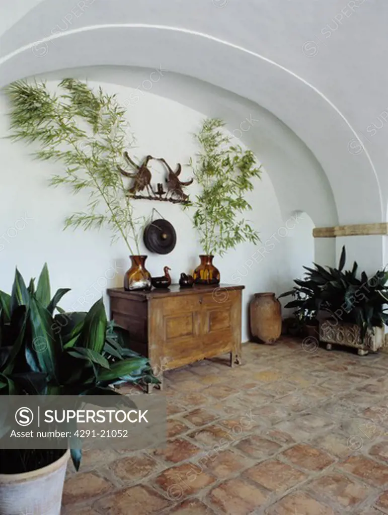 Terracotta tiled floor in Spanish hall with antique carved chest in alcove with green houseplants