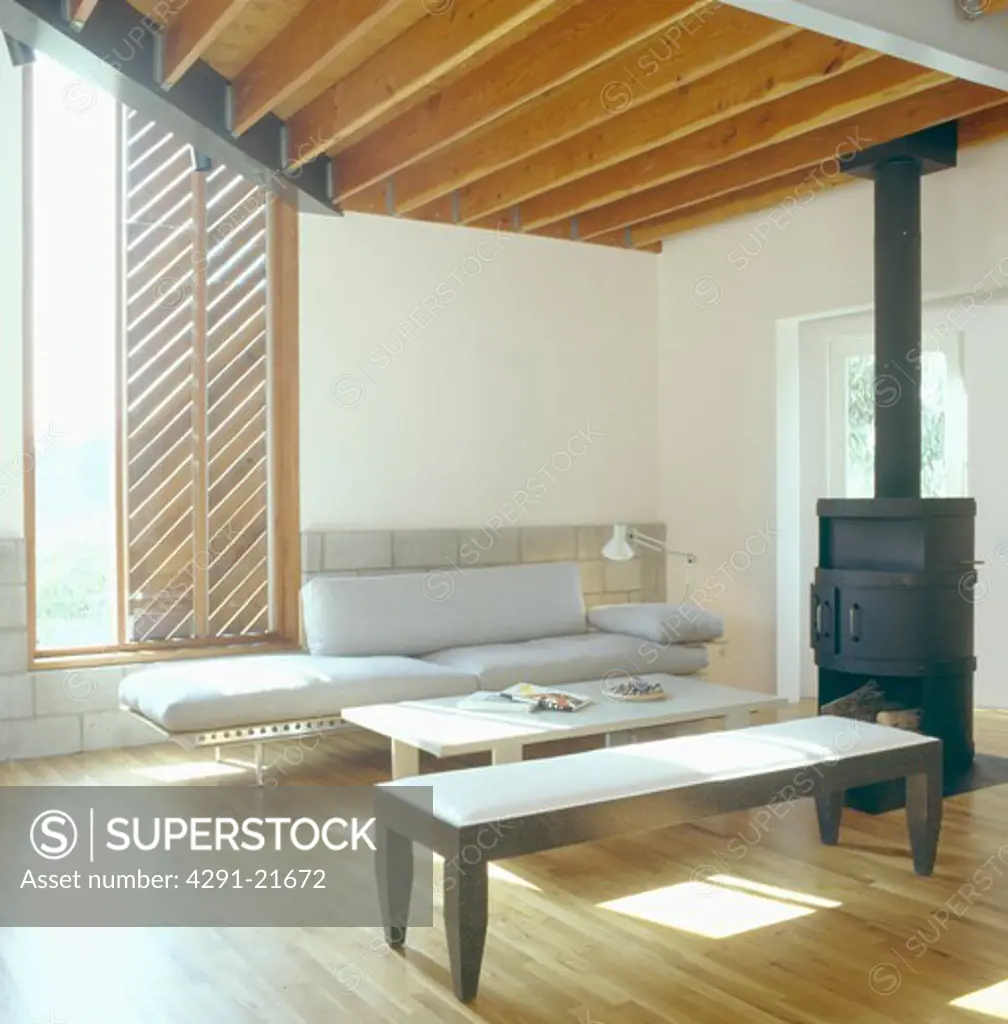 Modern stool and grey couch in architectural living room with black iron stove and smooth wood ceiling beams