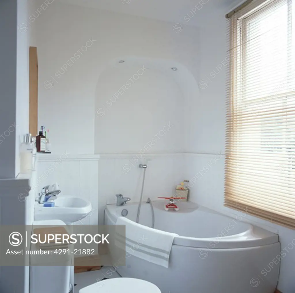 Slatted wooden blind on window above corner bath in modern white bathroom