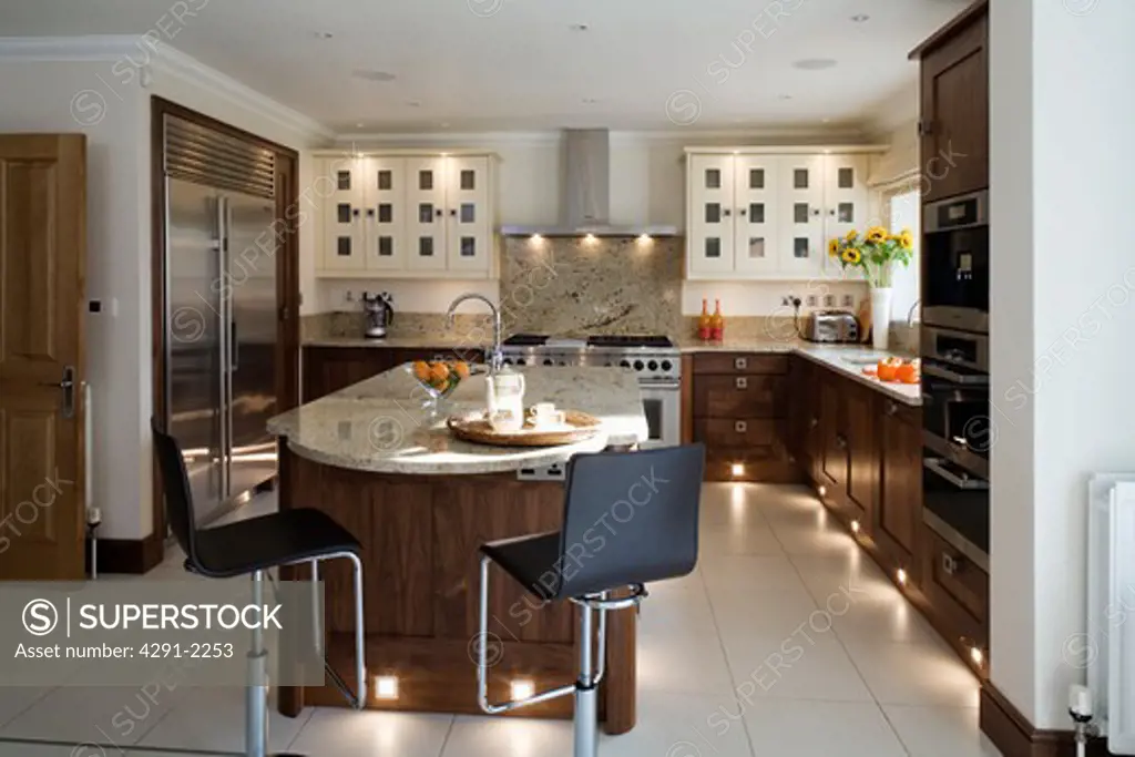 Chrome and black stools at island unit in modern kitchen with lighting in kickplates and white ceramic tiled flo