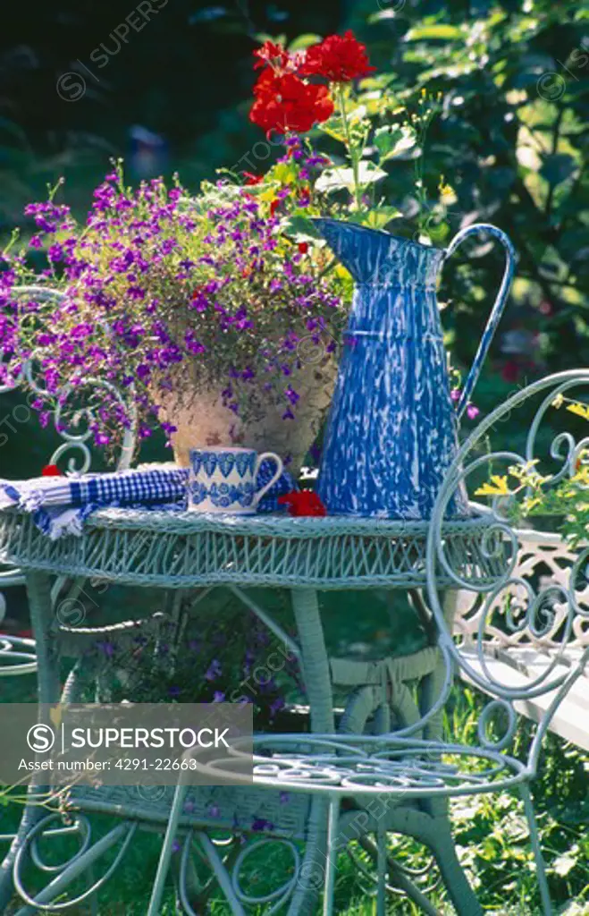 Tall blue and white enamel jug and blue lobelia in pot on blue wicker table with metal chairs in summer garden