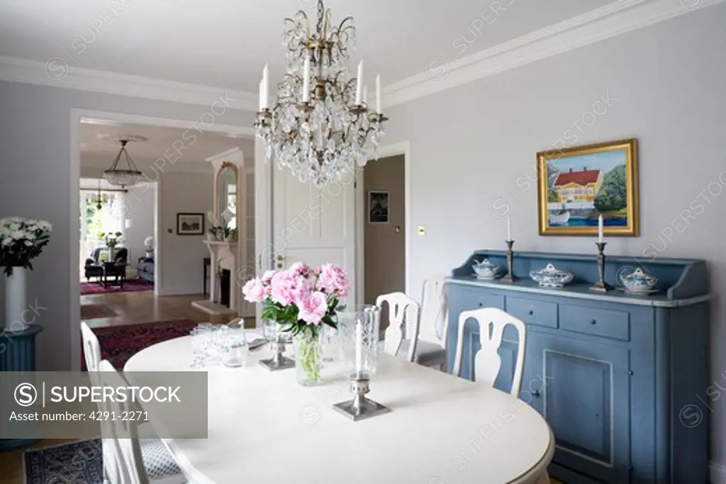 Glass chandelier above white painted table and chairs in Gustavian style dining room with antique Swedish sideboard