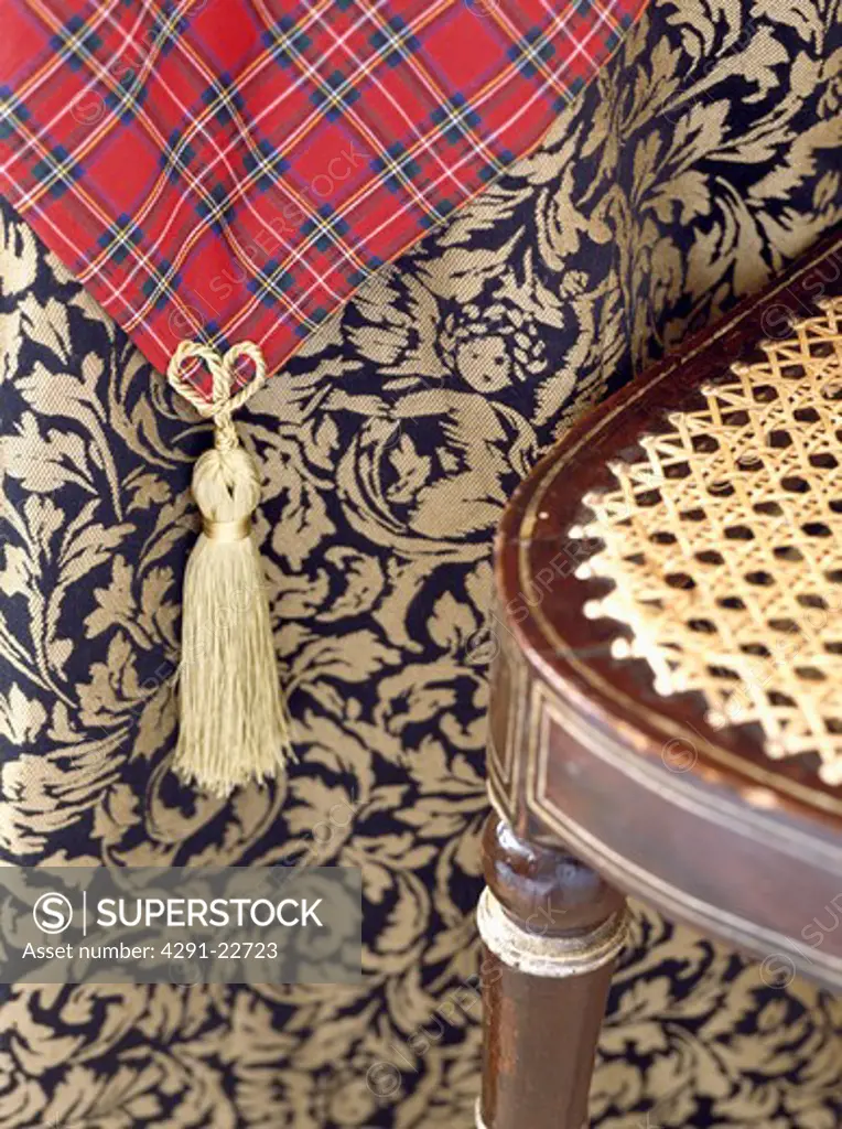 Close-up of cane-seated chair gainst patterned fabric with cream tassel on red tartan fabric