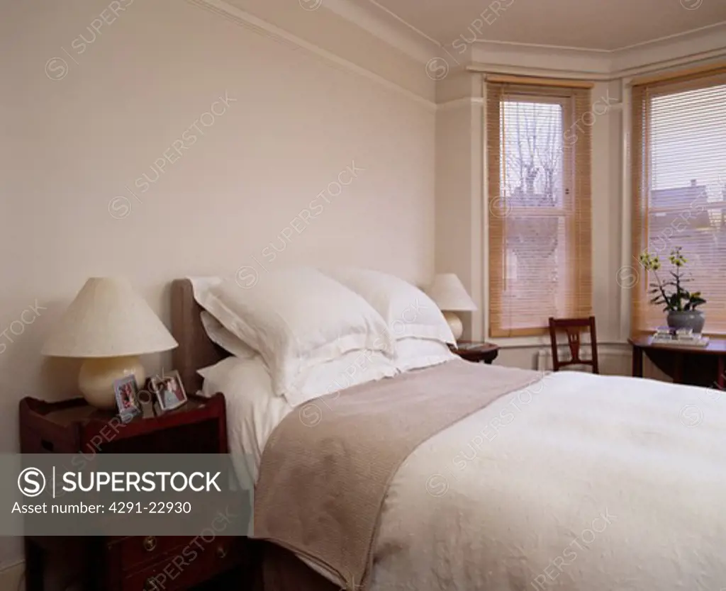 White pillows and linen on bed in cream bedroom with slatted wooden blinds on bay window