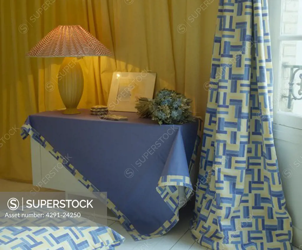 Lighted lamp on table with blue cloth edged with patterned fabric and matching curtains in town bedoom