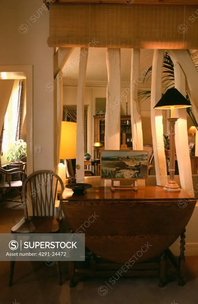 Lighted lamp on table in front of wall with white exposed beams below split-cane blind