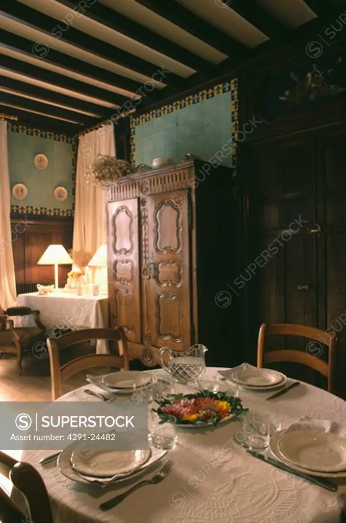 Place settings on white table cloth in dark green dining room with large antique cupboard and beamed ceiling