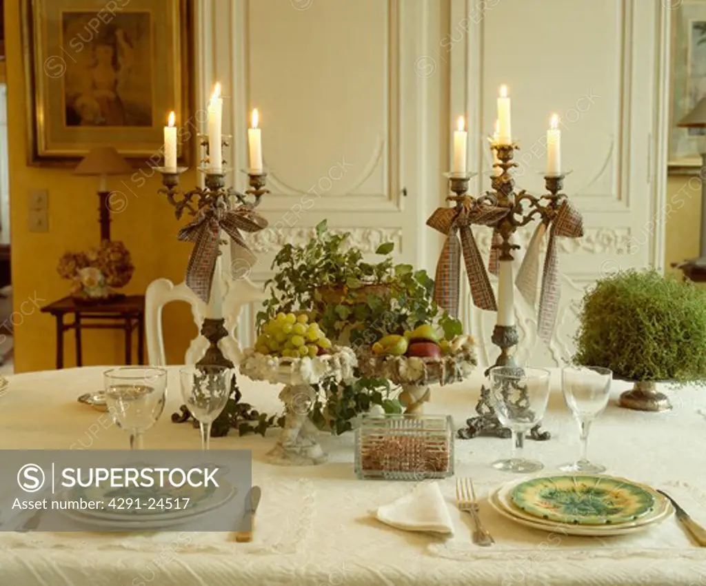 Table with wineglasses and plates with candles in tall candelabra and fruit decoration on white tablecloth