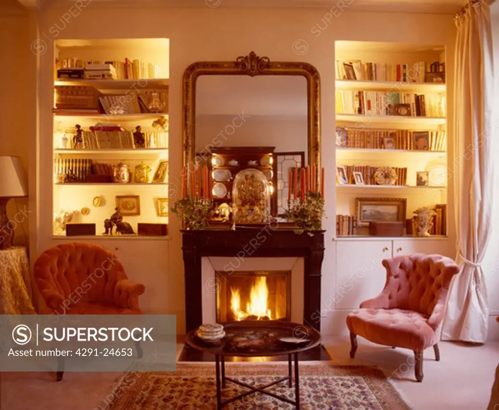 Lighted alcove shelves on either side of fireplace with lighted fire in living room with pink velvet chairs