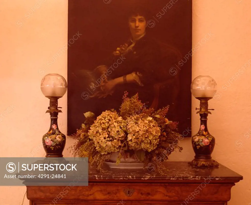 Close-up of oil portrait above Victorian glass lamps and dried flower arrangement