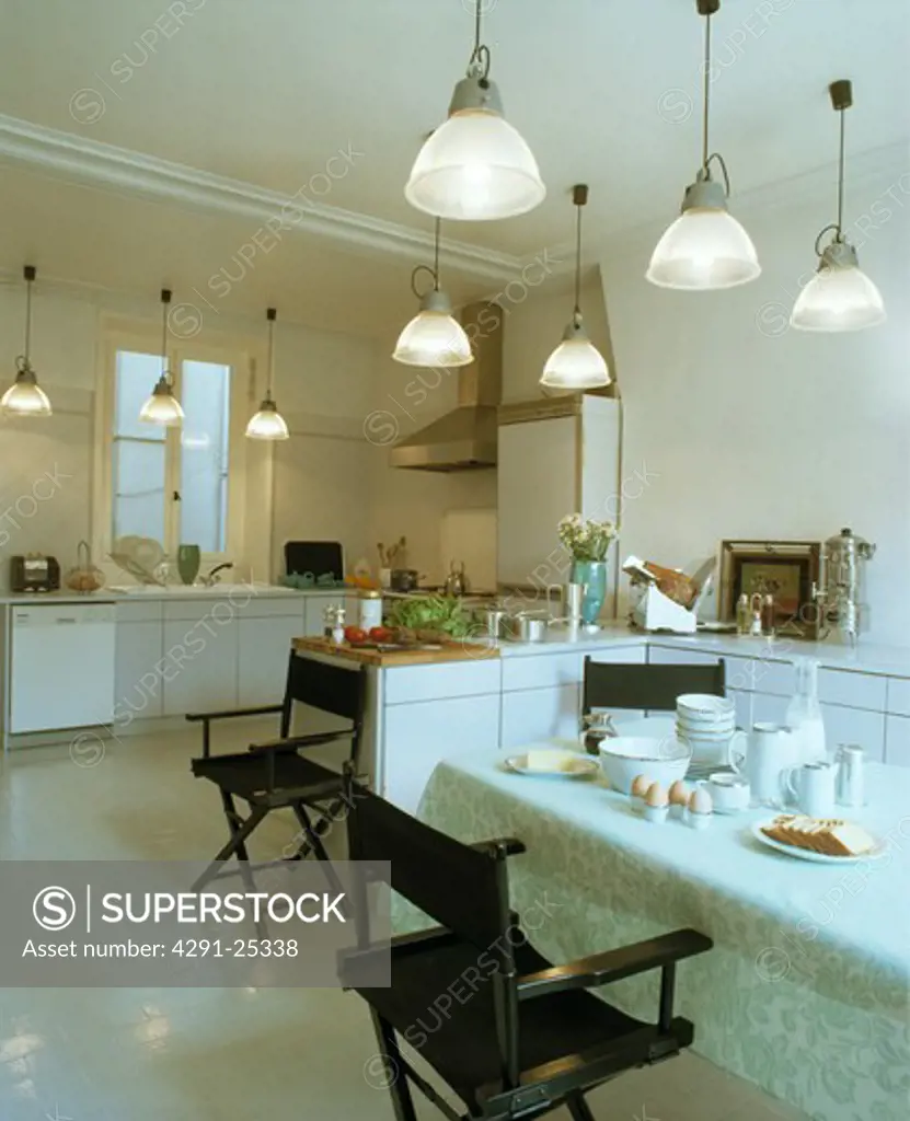 Pendant lights with glass shades in modern white kitchen with black director's chairs at table with white crockery
