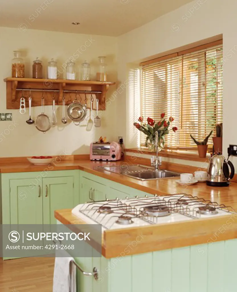 Gas hob in wooden worktop on peninsular unit in pastel green cottage kitchen with slatted wood blind