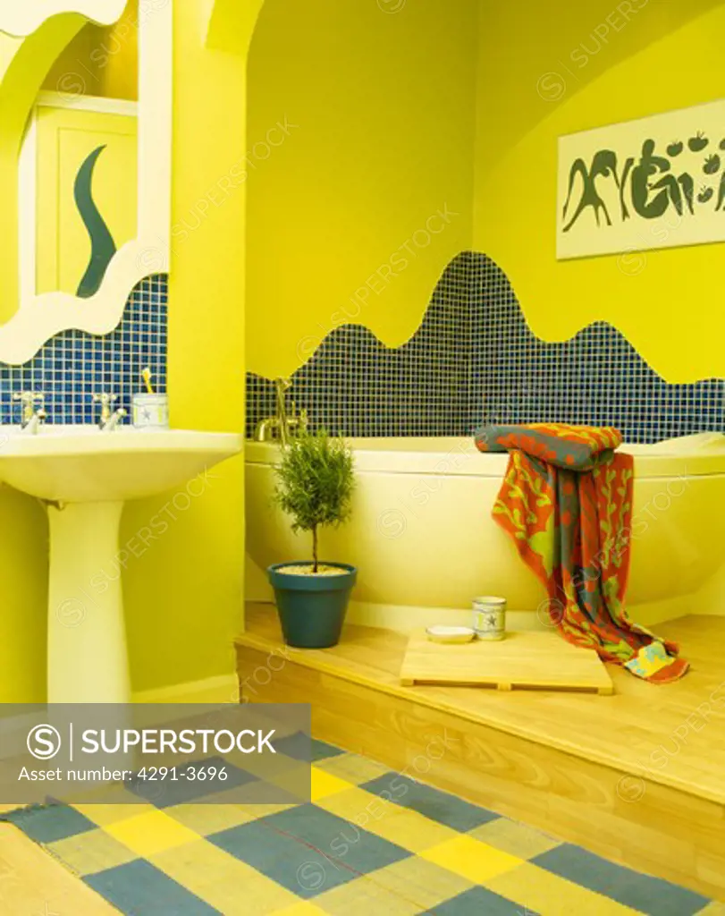 Mosaic tiling above corner bath in yellow split-level bathroom with yellow and blue checked rug below pedestal basin