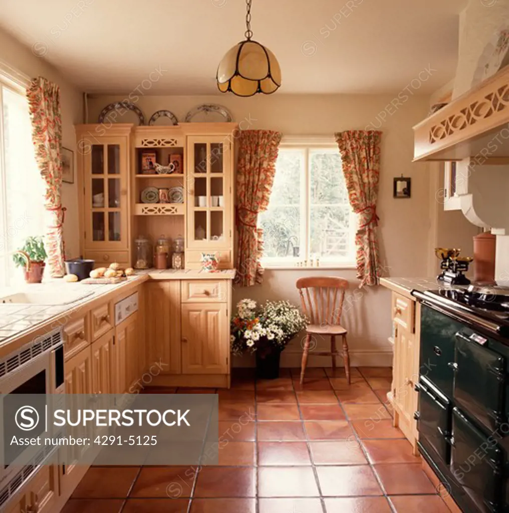 Terracotta floor tiles and black Aga in cottage kitchen with floral curtains and wooden chair