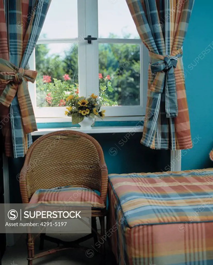 Wicker chair in front of window with blue and pink checked curtains in bedroom with matching checked bedcover