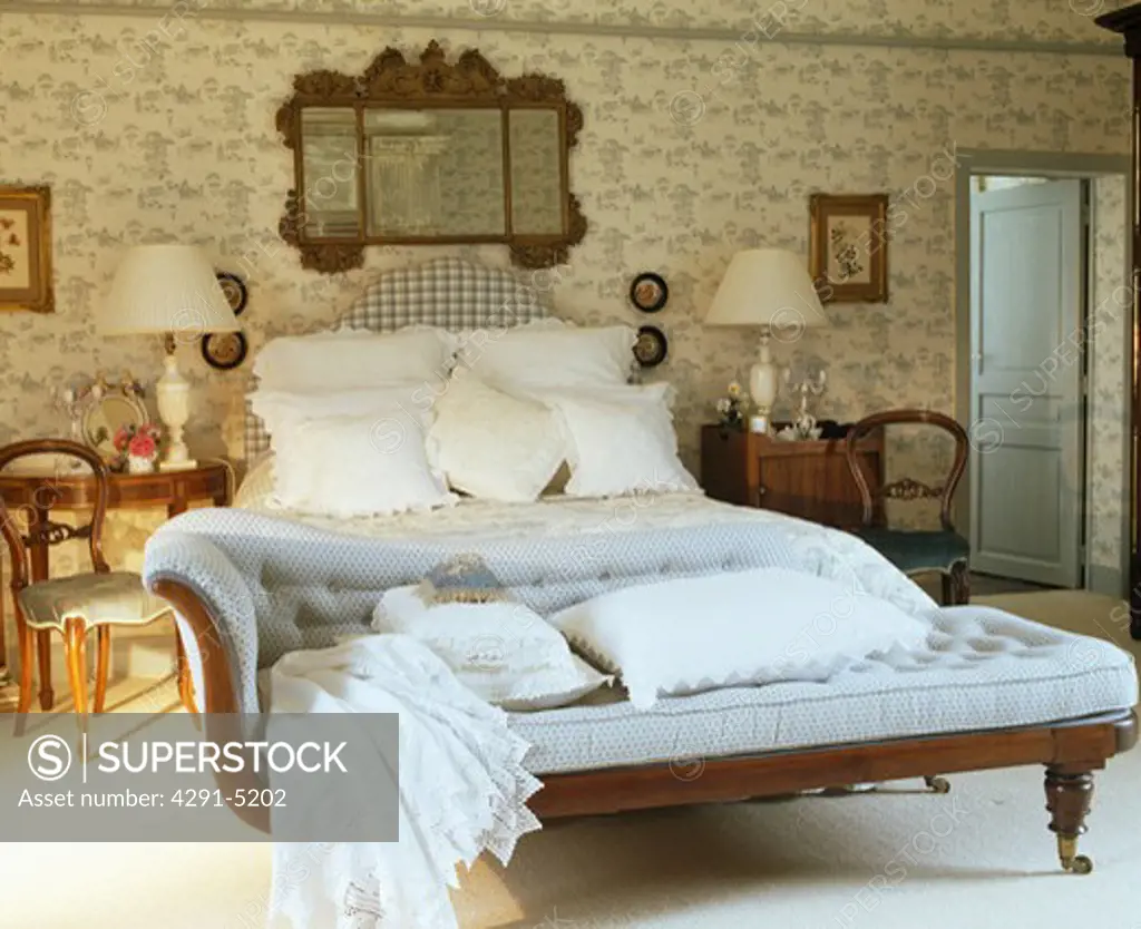 Lace-trimmed cushions and linen on pale blue chaise-longue below bed piled with white pillows in country bedroom