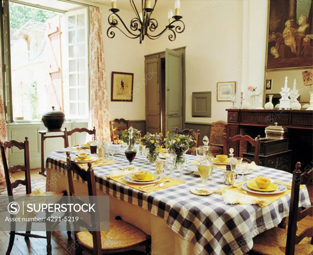 Yellow crockery on blue and white checked tablecloth in French country dining room with ladderback rush-seated chairs