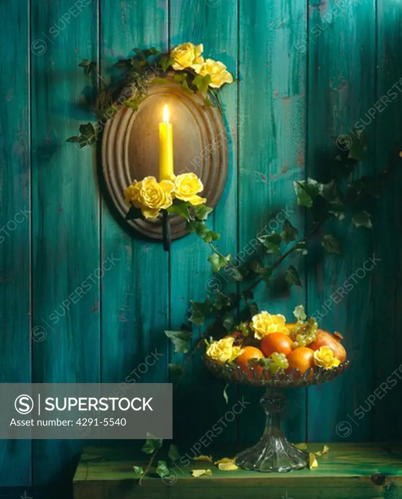 Yellow roses on mirror with lighted candle on green painted wooden wall above table with bowl of fruit and roses