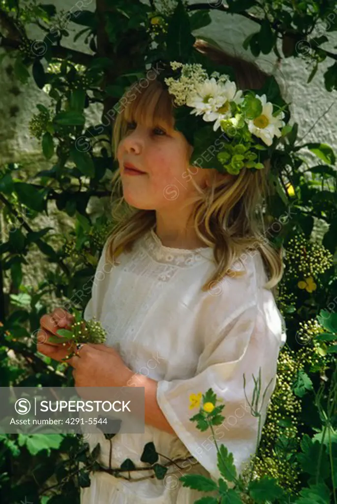 Close-up of small bridesmaid wearing coronet of daisies and simple white dress and standing among green leaves